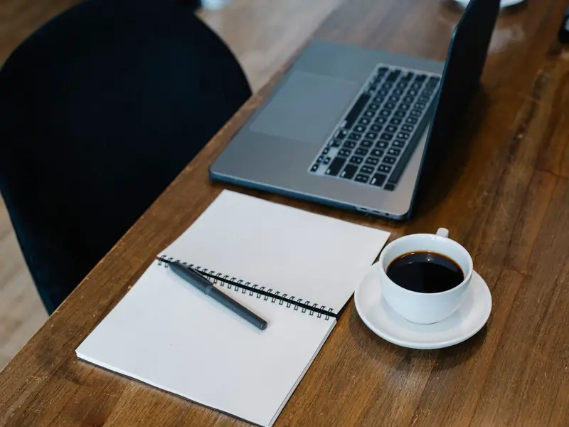A laptop, a coffee cup and an open spiral notebook with a pen on a wooden desk